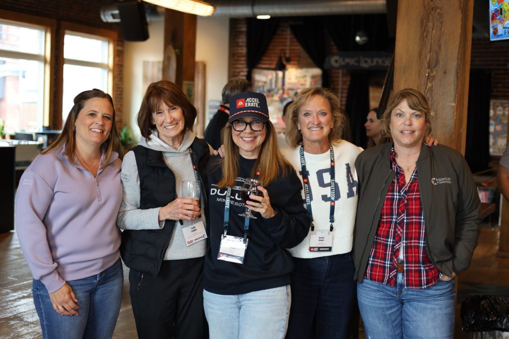 Five women smiling and posing together indoors at the ASB Accelerate Event in Duluth MN, wearing badge lanyards. One woman in the center holds a camera and wears a cap, while the others stand close on either side. Excitement for 2026 fills the air.