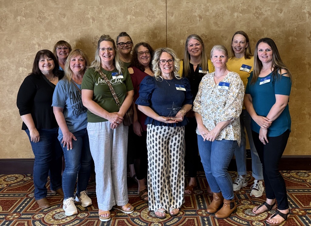 Twelve women stand together indoors in front of a beige wall, smiling at the camera. Most wear casual outfits, some have name tags. Lori Rogge holds a glass award, celebrating the Sales Support team at American Solutions for Business. The group stands on a patterned carpet.