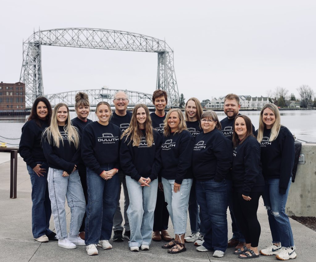 A group of 14 people wearing matching black “Duluth” sweatshirts pose together outdoors in front of the Aerial Lift Bridge in Duluth MN, with water and buildings behind them on a cloudy day during the ASB Accelerate Event 2026.