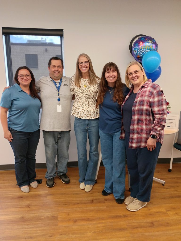Five smiling adults stand together indoors, posing for a group photo. There are balloons behind them, including one that says "Retirement." They are casually dressed and standing on a wood floor by a white wall.