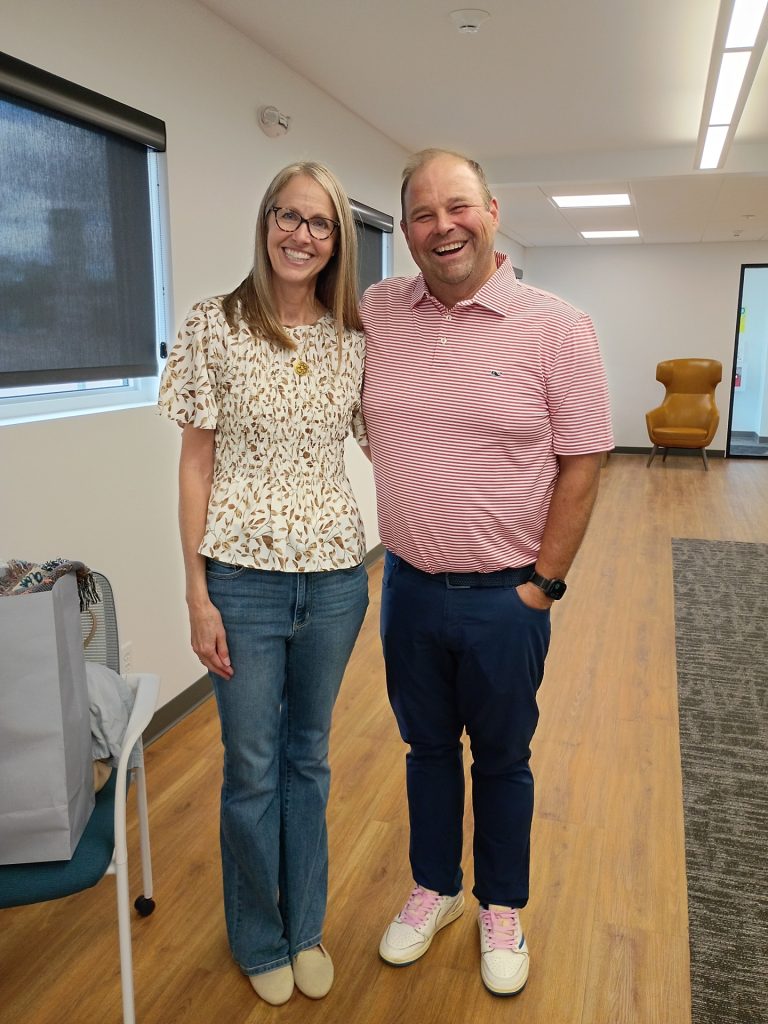 A smiling woman and man stand side by side in a modern room with wood flooring, windows, and a chair in the background. The woman wears glasses, a floral blouse, and jeans; the man wears a pink striped shirt and blue pants.