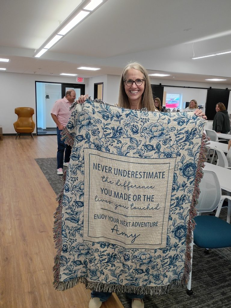 A smiling woman stands in an office holding a decorative blanket that reads, "NEVER UNDERESTIMATE the difference YOU MADE OR THE lives you touched. ENJOY YOUR NEXT ADVENTURE Amy." People are gathered in the background.