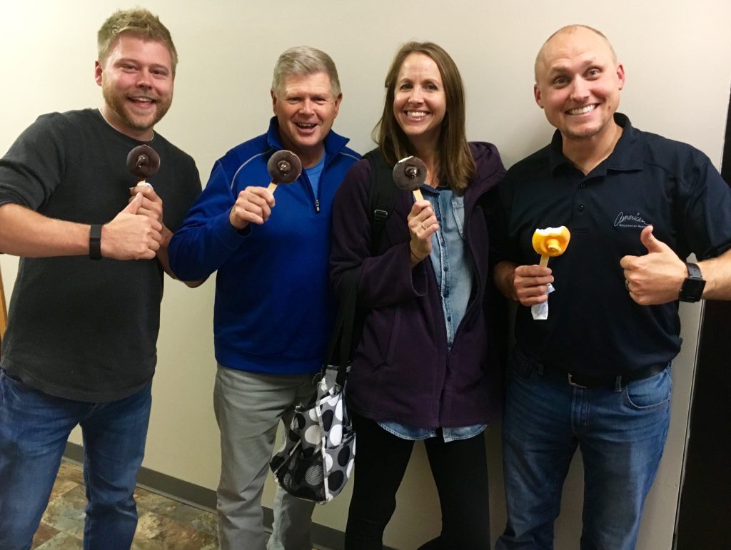 Four smiling adults stand indoors, each holding a different ice cream treat. Three hold chocolate-covered ice cream bars, while one holds an orange popsicle. They are casually dressed and appear cheerful.