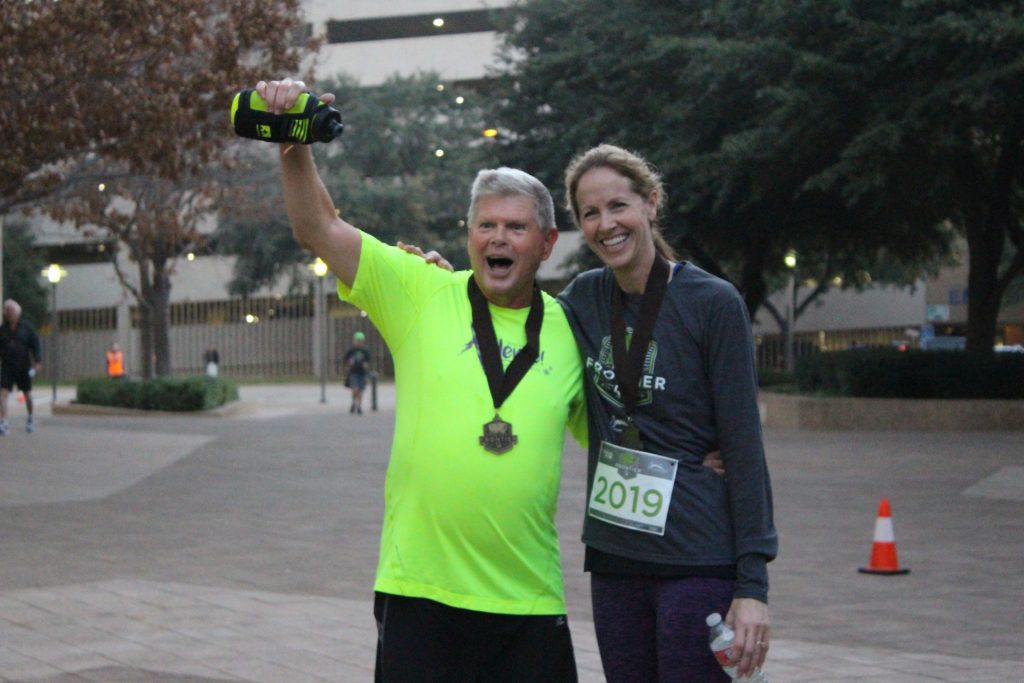Two marathon runners, wearing medals, smile and pose together after finishing a race. The man in a bright yellow shirt holds up a water bottle, while the woman in gray holds her own bottle. Trees and a building are in the background.