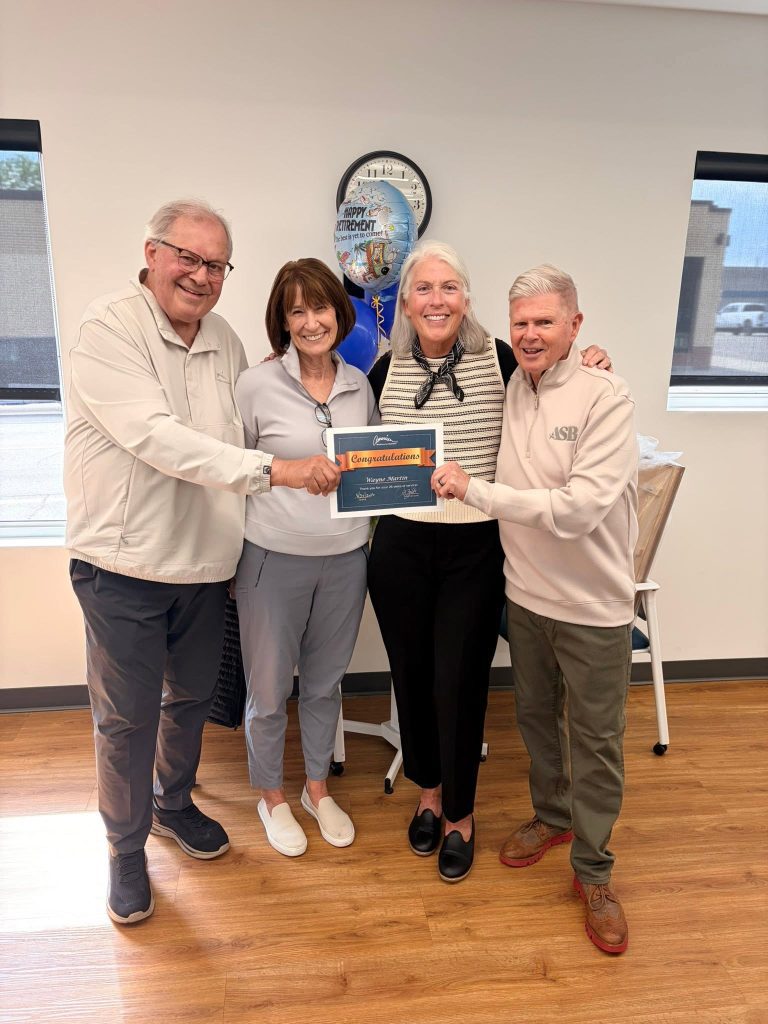 Four smiling adults stand in an office, holding a "Congratulations" sign together. A retirement-themed clock and blue balloon are in the background. They are all casually dressed and appear happy, celebrating an occasion.