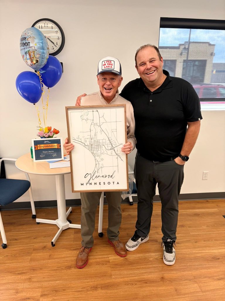 Two smiling men stand indoors; one holds a framed map labeled "Glenwood, Minnesota." Behind them is a table with a bouquet, balloons, a clock, and a window showing part of a building outside.