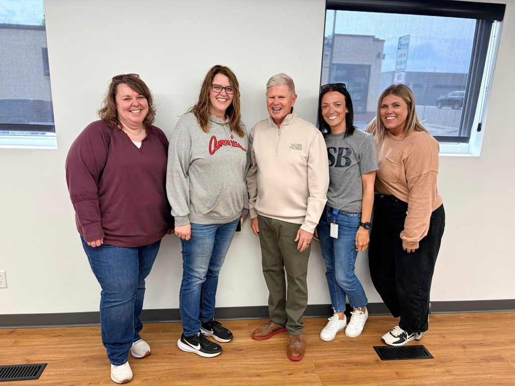 Five people stand smiling in a room with white walls and large windows. Four women stand on either side of an older man in the middle. They are all dressed casually and appear happy.